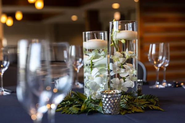 A centerpiece at a wedding with wine glasses on the table, flowers and floating candles in pillar vases, and blurred out lights in the distance.