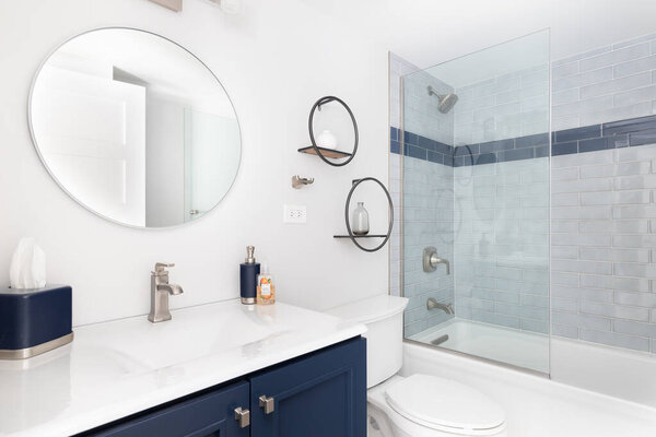 ELMHURST, IL, USA - JANUARY 20, 2021: A bright white bathroom with a blue vanity cabinet, white granite countertop, circular mirror, and a blue subway tile shower.