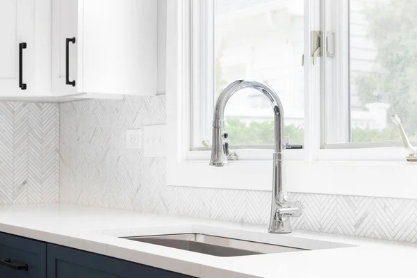 A kitchen sink detail shot with blue and white cabinets, herringbone tile backsplash, and a chrome faucet in front of a window.