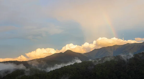 A sunset with a rainbow and large puffy clouds in the Great Smoky Mountain National Park. Smoke lingers in the mountains after a heavy storm.