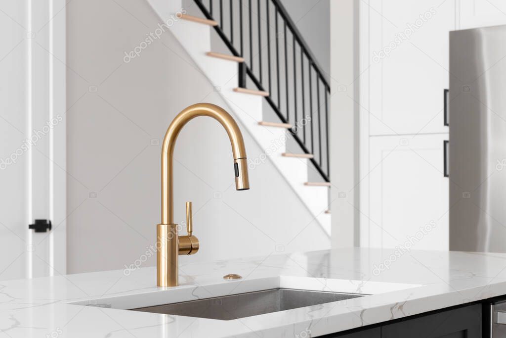 A kitchen sink detail shot with a gold faucet, grey island, white marble countertop, and lights hanging above the island.