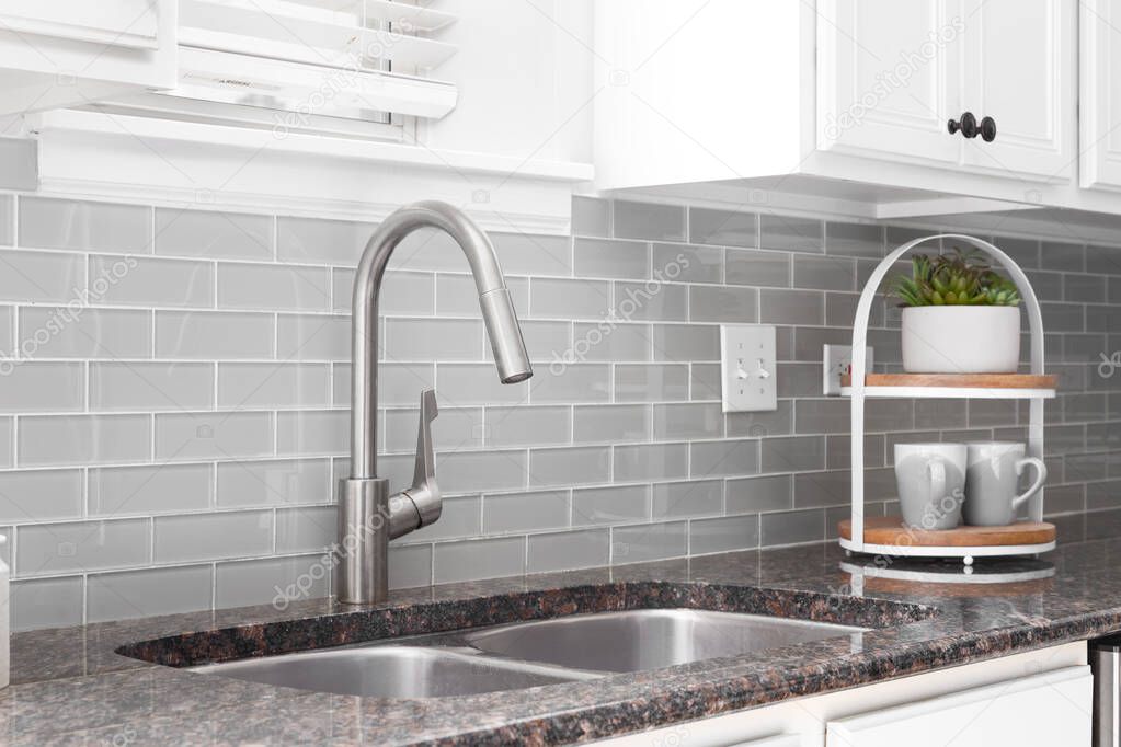 A kitchen sink detail shot with white cabinets, grey subway tile backsplash, and a granite countertop.