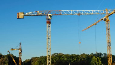 Şehir merkezindeki inşaat alanının panoramik manzarası. Mavi arka planda ağır inşaat malzemelerini kaldırmak için birkaç uzun sarı vinç var. İnşaat endüstrisi. Metin için boşluk.