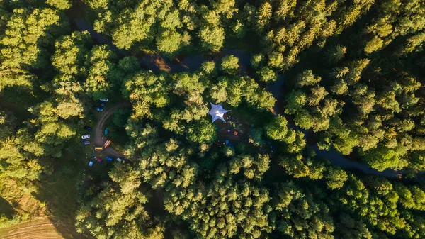 Aerial drone view of big tent between trees in evening forest on riverbank.