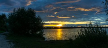 Gölde dramatik bir gün batımı. Gölün kıyısında gün batımı. Yansıma. Mavi gökyüzü ve sarı güneş ışığı. Gün batımında manzara. Panoramik pankart. Doğa konsepti.