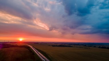 Kırsal kesime doğru akşam güneşiyle giden güzel, dolambaçlı bir yol. Dramatik gökyüzü arkaplanı. Kırmızı günbatımında otobanın havadan görünüşü. Ulaşım geçmişi. Kırsal alanlara yakın yolu olan manzara.