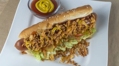 Marullu sosisli, turşulu, kızarmış soğanlı, beyaz tabakta ketçaplı. Danimarka malzemeleriyle hazırlanmış sosisli sandviç. Fast food kavramları.