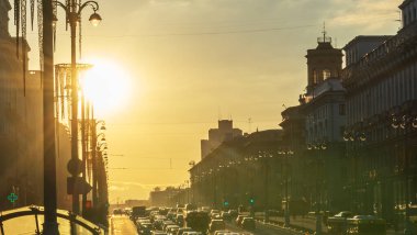 Şehirdeki binalar arasında günbatımının panoramik görüntüsü. Ilık bir yaz akşamında yol üzerinde parlak sarı bir güneş ışığı. Yolun sonunda, güneş ufuktan batar. Arabaların ön planında..