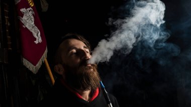 Büyük sakallı ve şık saç kesimli vahşi bir adam koyu arka planda elektronik sigara ya da nargile içiyor. Buhar, buhar, nargile.