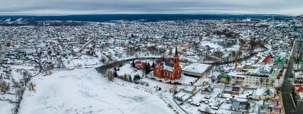 Kızıl Katolik kilisesi ve kışın küçük bir kasabanın havadan görünüşü. Karla kaplı küçük bir Avrupa kasabası. Kilise, evler, nehir ve nehir kenarları olan cennet manzarası. Din kavramları.