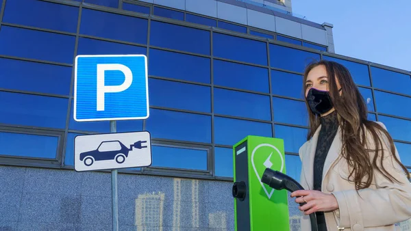 Elektrikli, hibrit arabaları şarj etmek için kolonun yanında şarj kablosu olan koruyucu tıbbi maskeli kadın. Trafik levhası, park etme ve elektrikli araçları şarj etme. Coronavirus ve ekoloji kavramı.