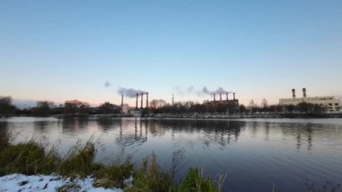 Şehrin ortasında suyun yanında hava kirliliğine yol açan fabrikanın hızlandırılmış videosu. Kışın termik santraller. Ekoloji kirliliği. Fabrika boruları kalın beyaz duman buharı.
