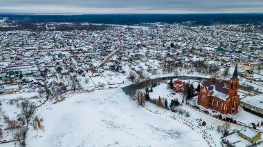 Kızıl Katolik kilisesi ve kışın küçük bir kasabanın havadan görünüşü. Karla kaplı küçük bir Avrupa kasabası. Kilise, evler, nehir ve nehir kenarları olan cennet manzarası. Din kavramları.