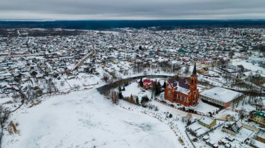 Kızıl Katolik kilisesi ve kışın küçük bir kasabanın havadan görünüşü. Karla kaplı küçük bir Avrupa kasabası. Kilise, evler, nehir ve nehir kenarları olan cennet manzarası. Din kavramları.