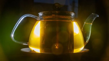 Karanlık arka planda mercek alevi olan çaydanlığın yakın görüntüsü. Sıcak yeşil çayla parıldayan küçük cam çaydanlık. Mesaj için yer var. Çay seremonisi için..