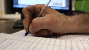 Arka planda bilgisayarı olan ve bir fabrikada yer alan bir defterde notlar alan bir adamın eli. 