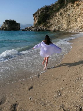 Genç, sıska, beyaz gömlekli, deniz kenarında koşan ve arka planda kayalıklar olan kumlu kumsalda eğlenen güzel bir kadın.