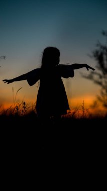 Çimenlerde dans eden ve gün batımını izleyen sevimli küçük bir kızın silüeti. Yüksek kalite fotoğraf
