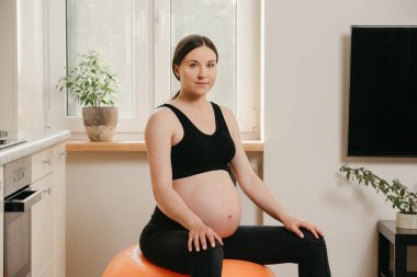 Hamile bir kadının portresi. Turuncu bir egzersiz topunun üzerinde oturuyor ve evindeki mutfağın yakınındaki bir spor salonundan sonra dinleniyor. Sağlıklı hamilelik.