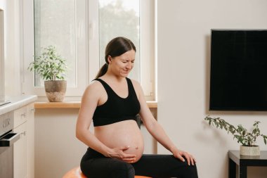Evinin mutfağının yanındaki turuncu egzersiz topunun üzerinde kolu karnında acı içinde kıvranan hamile bir kadının portresi. Karmaşık hamilelik. 