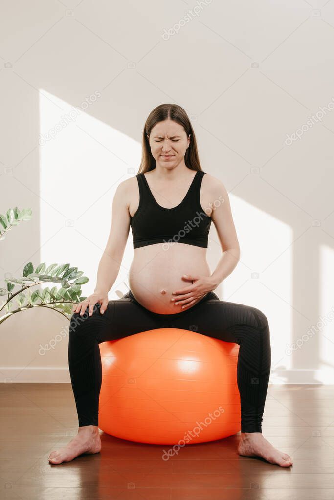 A pregnant woman is writhing in pain with her arm on her belly on an orange exercise ball at her home in the evening. Complicated pregnancy.