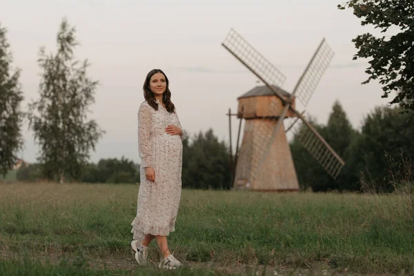 風車の中で妊娠中の女性が農場を歩いている間 彼女の腹を手で握っている 幸せな妊娠だ 夜の牧草地で長いドレスを着た女性 — ストック写真