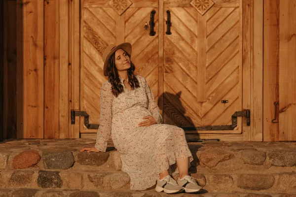 妊娠中の女性が東ヨーロッパの村の古い教会の石段でリラックスしています 幸せな妊娠だ 長いドレスを着た女性と夜に田舎に座っている帽子 — ストック写真