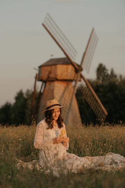 風車の背景にいる妊婦は草の上で冷えていて 夕方に牧草地で小瓶を持っています 農場で幸せな妊娠 長いドレスを着た女性と藁帽子 — ストック写真