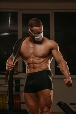 Spor yaptıktan sonra poz veren, tıbbi maske takan üstsüz bir vücutçunun orta boy bir pozu. Cerrahi maskeli kaslı bir adam spor salonunda kaslarını geriyor..