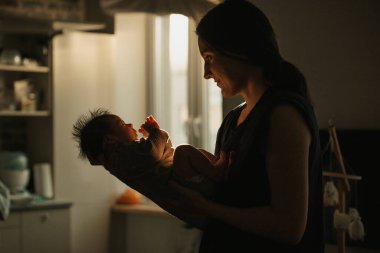 Yeni doğmuş bebeği kucağında tutan annenin orta uzunlukta bir görüntüsü. Bebek ve anne gün ışığında pencerenin önündeler..