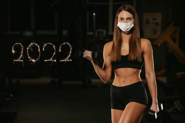 A fit woman in a face mask to avoid the spread of coronavirus is doing bicep curls with dumbbells. A sporty girl in a surgical mask is posing during the arms workout in the gym. 2022