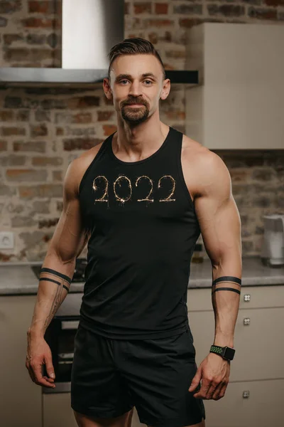 一个长着胡子的善良而强壮的男人正在他的公寓里做运动后的姿势 一个手臂上有纹身的运动员正在家里展示他的衬衫和2022年 一个聪明的人在手腕上 — 图库照片