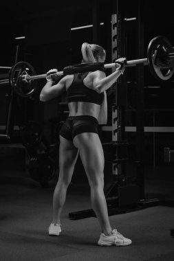 Spor salonundaki çömelme yerinin yanındaki halterle çömelmeye başlayan sarı saçlı, sportif bir kadının fotoğrafı. Bir kız bacak egzersizi yapıyor..