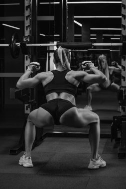 Spor salonundaki çömelme yerinin yanında halterle çömelmeden önce ısınan sarı saçlı, sportif bir kadının fotoğrafı. Bir kız bacak egzersizi yapıyor..