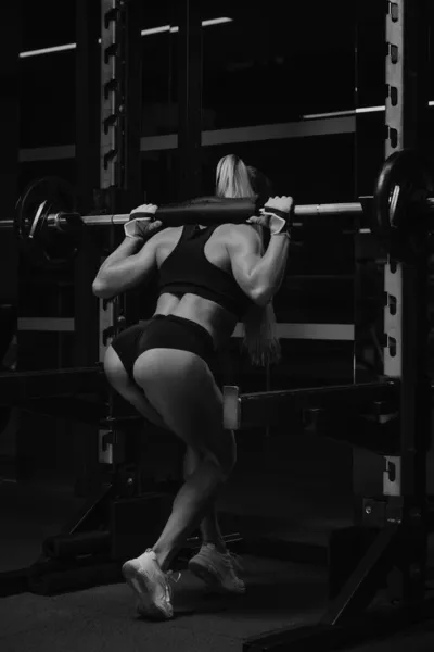 一个金发碧眼的健康女人在体育馆的蹲架上蹲下后 正在把杠铃放下 一个女孩在做腿部锻炼 — 图库照片