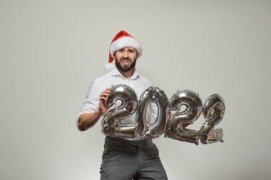 Kırmızı kadife Noel Baba şapkalı mutsuz bir adam elinde 2022 şeklinde gümüş balonlar tutuyor. Yeni yıl partisinde sakallı bir adam.