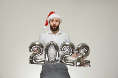 Kırmızı kadife Noel Baba şapkalı sürpriz bir adam elinde 2022 şeklinde gümüş balonlar tutuyor. Yeni yıl partisinde sakallı mutlu bir adam..