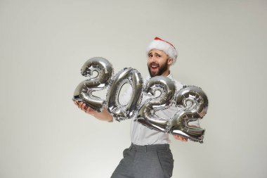 Kırmızı kadife Noel Baba şapkalı şık bir adam elinde 2022 şeklinde gümüş balonlar tutuyor. Yeni yıl partisinde sakallı, gülümseyen bir adam..