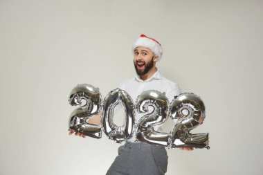 Kırmızı kadife Noel Baba şapkalı mutlu bir adam 2022 'de gümüş balonlar gösteriyor. Yeni yıl partisinde sakallı, gülümseyen bir adam..