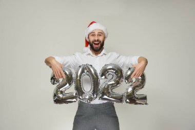 Kırmızı kadife Noel Baba şapkalı mutlu bir adam 2022 'de gümüş balon kaldırıyor. Yeni yıl partisinde sakallı, mutlu bir adam..