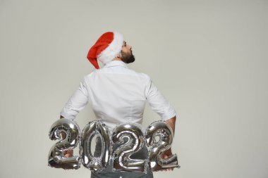 Kırmızı kadife Noel Baba şapkalı bir adam sırtındaki gümüş balonları 2022 şeklinde kaldırıyor. Yeni yıl partisinde sakallı bir adam başını sağa çevirdi..
