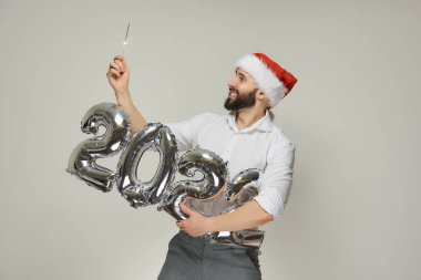 Kırmızı kadife Noel Baba şapkalı gülümseyen bir adam bir elinde 2022 şeklinde gümüş balonlar diğer elinde ise maytap tutuyor. Sakallı bir adam yılbaşı partisinde maytaplara bakıyor..