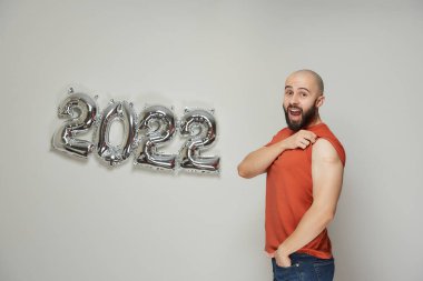 Sakallı kel bir adam 2022 'deki gümüş balonların yanında aşıdan sonra omzunda tıbbi bir yara bandı gösteriyor.. 