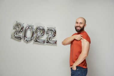 Sakallı mutlu kel bir adam gülümsüyor ve 2022 'de aşıdan sonra gümüş balonların yanında omzunda tıbbi bir yara bandı gösteriyor.. 