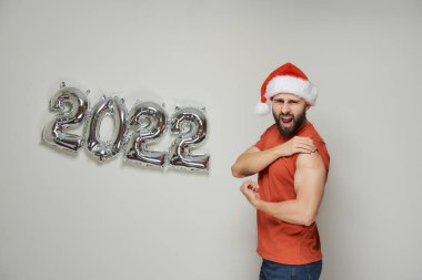 Noel Baba şapkalı, şaşırmış kel bir adam, Coronavirus (Covid-19) aşısından sonra, 2022 yılında durdurulmuş gümüş balonların yanında alçılı pazıları gösteriyor..