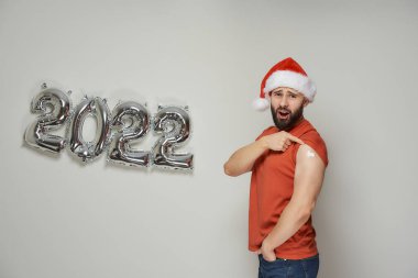 Noel Baba şapkalı kel bir adam, Coronavirus (Covid-19) aşısından sonra 2022 'de gümüş balonların yanında alçıyla omzunu gösteriyor..