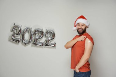 Noel Baba şapkalı kel bir adam, 2022 yılında Coronavirus (Covid-19) aşısından sonra gümüş balonların yanında gülümsüyor ve omzunu sıva ile gösteriyor.. 