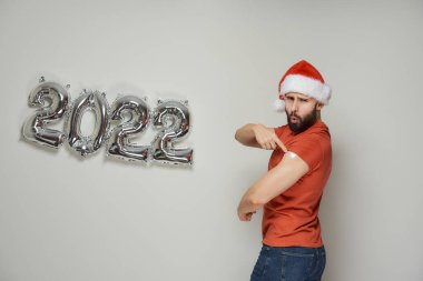 Noel Baba şapkalı, flört eden bir adam, Coronavirus (Covid-19) aşısından sonra, 2022 'de gümüş balon şeklinde bir omuz ve sıva gösteriyor.. 