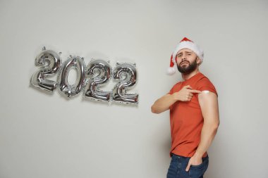 Noel Baba şapkalı ciddi bir adam, Coronavirus (Covid-19) aşısından sonra 2022 yılında gümüş balon şeklinde bir omuz ve sıva gösteriyor..