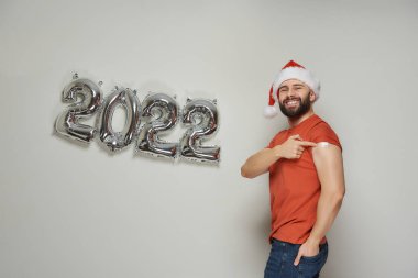 Noel Baba şapkalı mutlu bir adam, Coronavirus (Covid-19) aşısından sonra 2022 'de gümüş balonların yanında alçılı bir omuz gösteriyor..